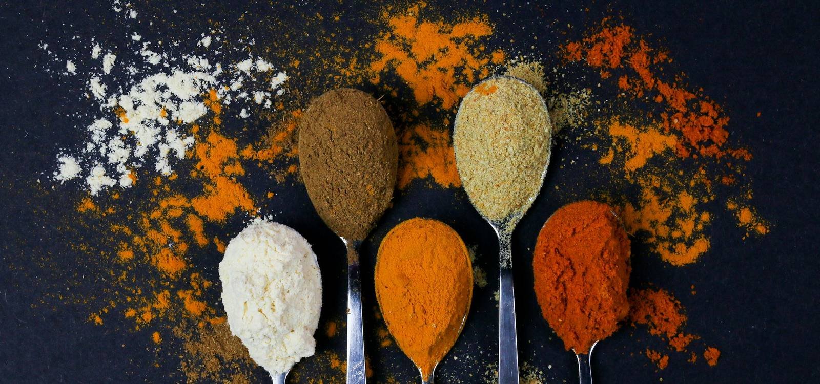 Ground spices on spoons scattered across a dark surface
