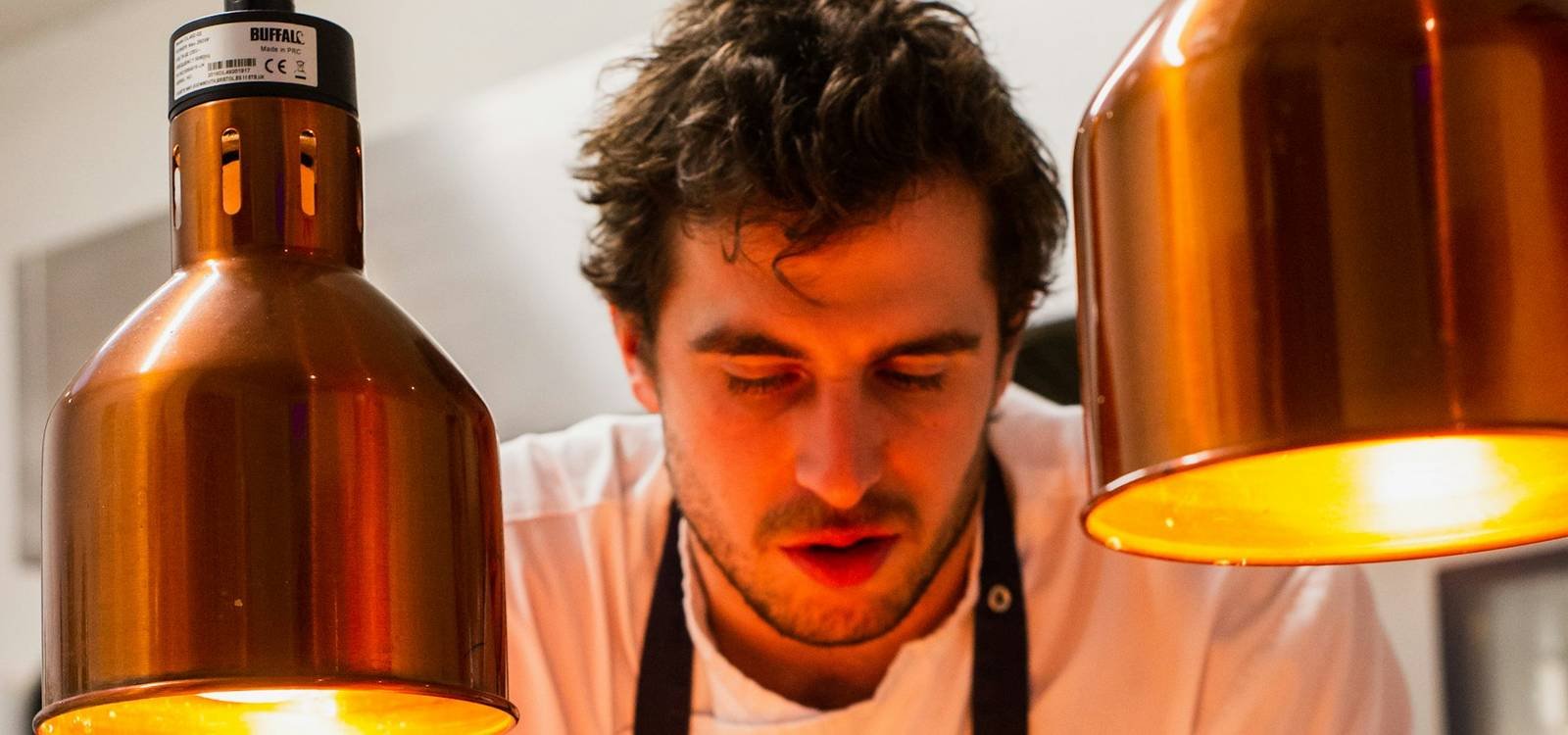 A chef plating a dish under copper pass lamps